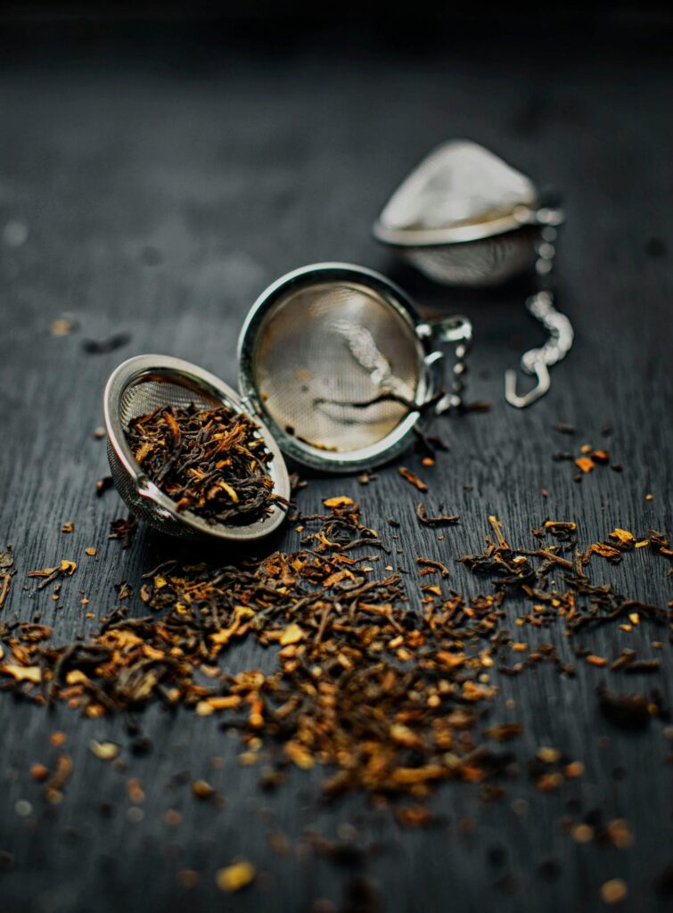 Loose dried tea leaves scattered with a metal tea strainer on a dark surface.